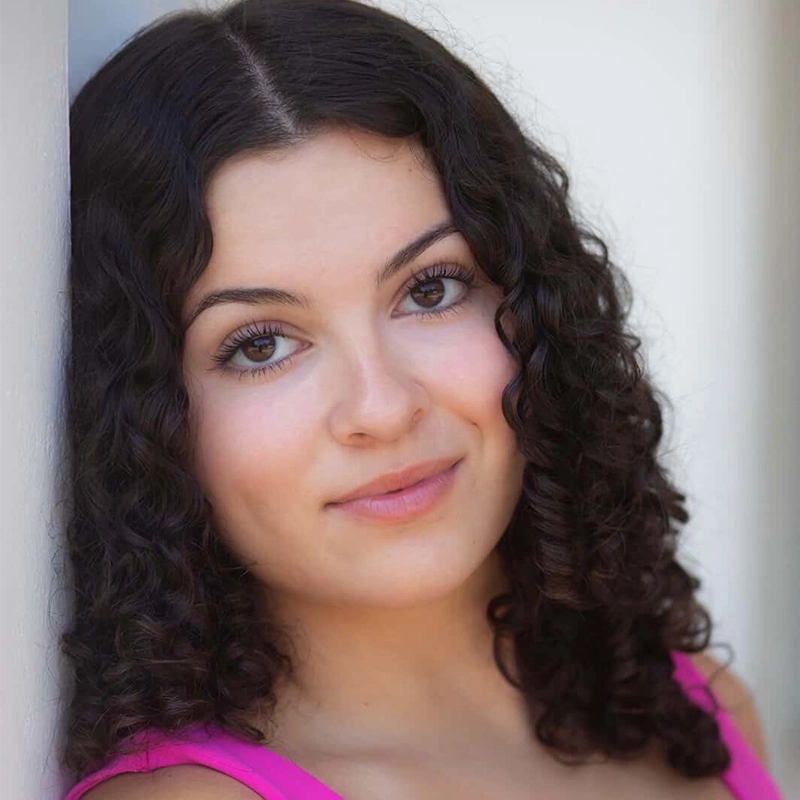 A young woman with medium skin, dark curly hair, and brown eyes smiles softly. She is wearing a bright pink top and leans her head gently against a white wall, the background blurred to highlight her face in this Josiah Jennings-inspired portrait.