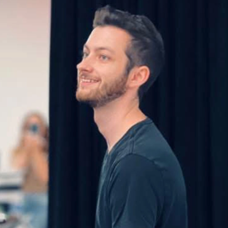 Evan Swanson, a man with short brown hair and a trimmed beard, wearing a dark t-shirt, smiles as he looks to his left. He is inside, in front of a black curtain; the blurred background shows another person holding a camera.