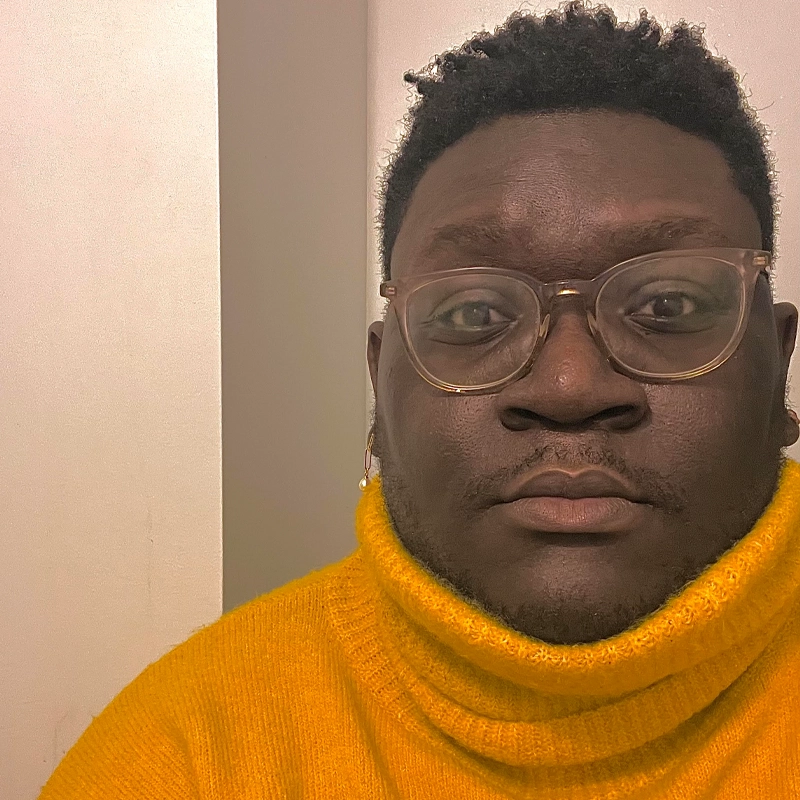 A person with dark skin, short curly hair, and clear round glasses looks directly at the camera. Ifeanyi Ezeanya wears a bright yellow turtleneck sweater. The background is plain, with off-white walls and a visible corner.