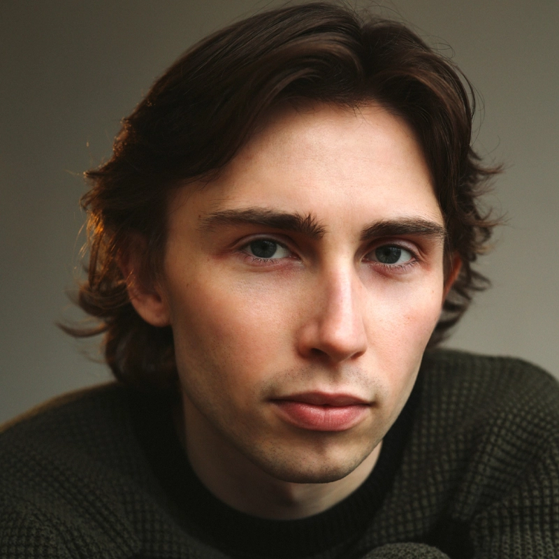A young person with light skin, medium-length brown hair, and blue eyes gazes directly at the camera with a neutral expression. Wearing a dark green textured sweater, Eddie Falshaw is set against a softly lit background in neutral tones.
