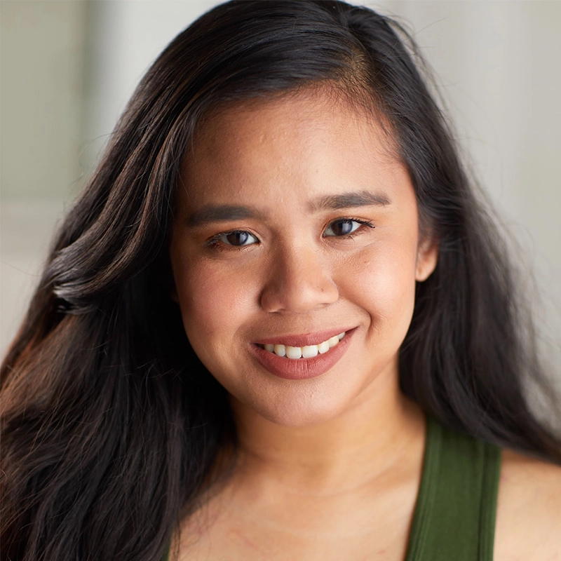 Fara Faidzan, a young woman with long, dark hair, smiles warmly at the camera. She is wearing a green tank top and natural makeup. The background is softly blurred, keeping the focus on her friendly expression.