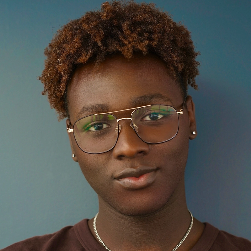 Ty Ford-Everett, a young person with short, curly brown hair and dark skin, looks at the camera. They wear large, gold-rimmed glasses, silver stud earrings, and a thin silver necklace against a plain blue wall with soft, even lighting.