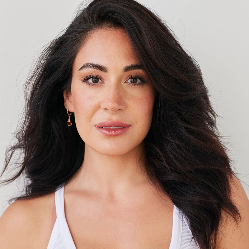 A woman with long, wavy dark hair and brown eyes looks at the camera. She wears a white tank top and small gold earrings. Her makeup is natural, and she stands against a plain off-white background, softly smiling.