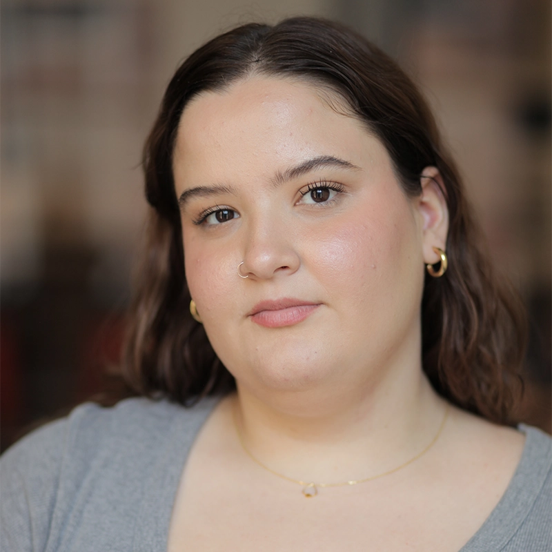 Phoebe Eskovitz, a woman with medium-length brown hair, gold hoop earrings, a gold necklace, and a grey top, looks directly at the camera with a neutral expression. The softly blurred background draws focus to her face.