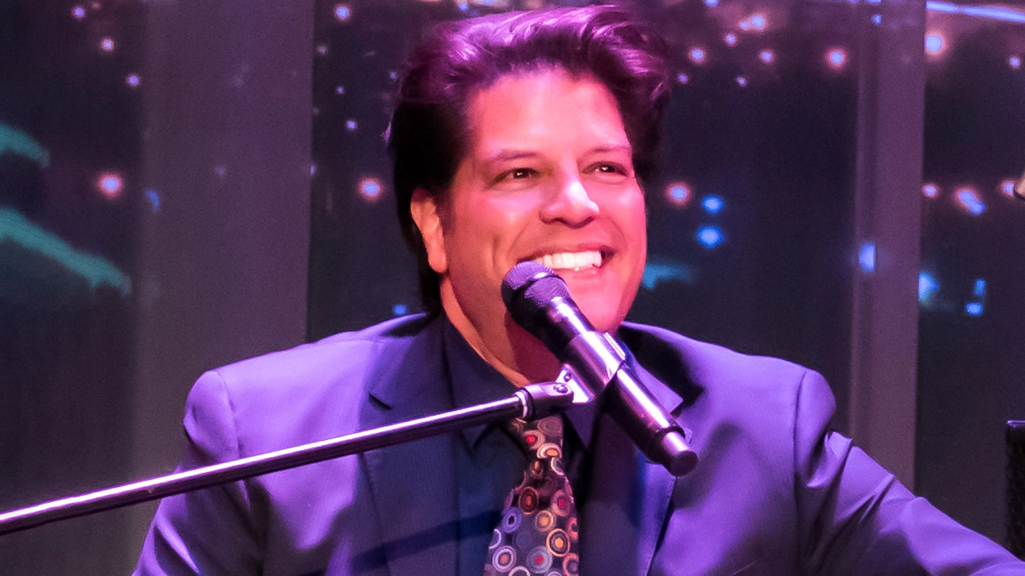 George Bugatti, in a dark suit, patterned tie, and blue shirt, smiles while singing or speaking into a microphone. Seated at a keyboard, he brings Broadway Jazz to life amid colorful blurred lights that hint at a vibrant stage performance.