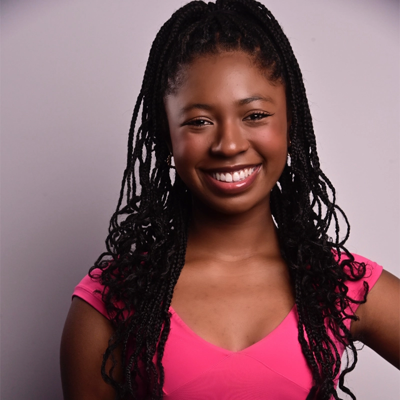 A young woman with long braided hair smiles at the camera against a plain light gray background. She is wearing a bright pink short-sleeved top and has one hand resting on her hip.