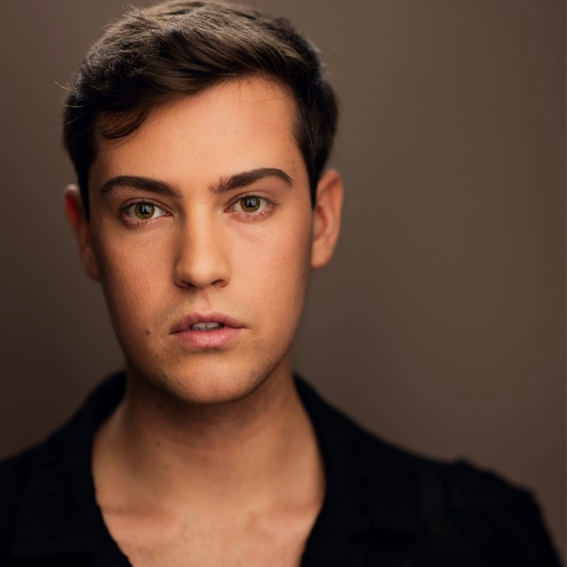 A young person with short brown hair and light skin gazes directly at the camera. With green eyes and groomed eyebrows, Blake Gioviti wears a black shirt with an open collar against a softly blurred, warm brown background.