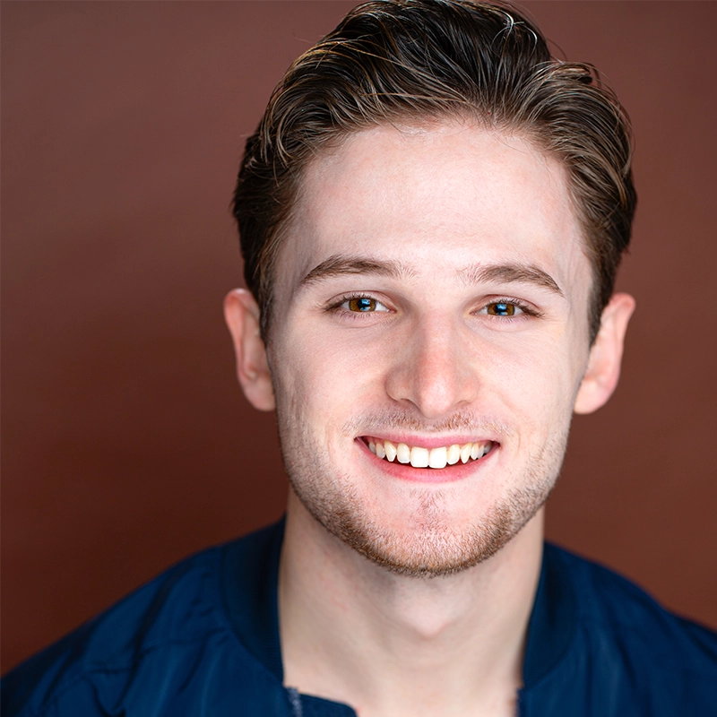 A young man with light skin, short brown hair, and blue eyes smiles at the camera. He has slight stubble and is wearing a dark blue jacket over a white shirt. The well-lit image features a plain brown background, photographed by Adriana Ellis.