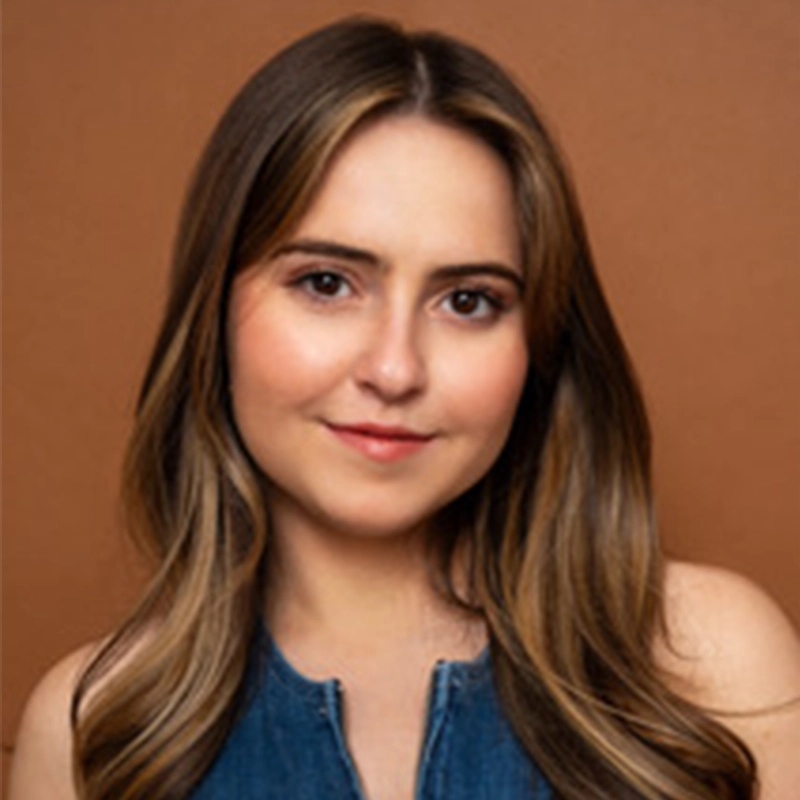 A young woman with long, straight, brown hair and fair skin smiles softly. Marguerite A. Boone is wearing a sleeveless blue denim top and is posed in front of a plain, warm brown background. The lighting is even, highlighting her facial features.