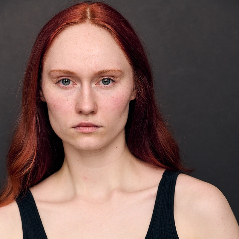 A young woman, Amanda Harris, with long, straight red hair and fair, freckled skin stares directly at the camera with a neutral expression. She wears a black sleeveless top and is set against a plain dark background.