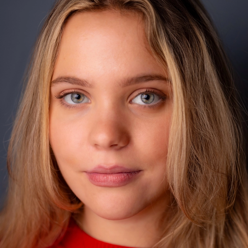 Samantha Hallenberg, a young woman with long blonde hair and light blue eyes, gazes directly at the camera. She has a neutral expression, subtle makeup, and wears a red top against a plain, dark grey background.