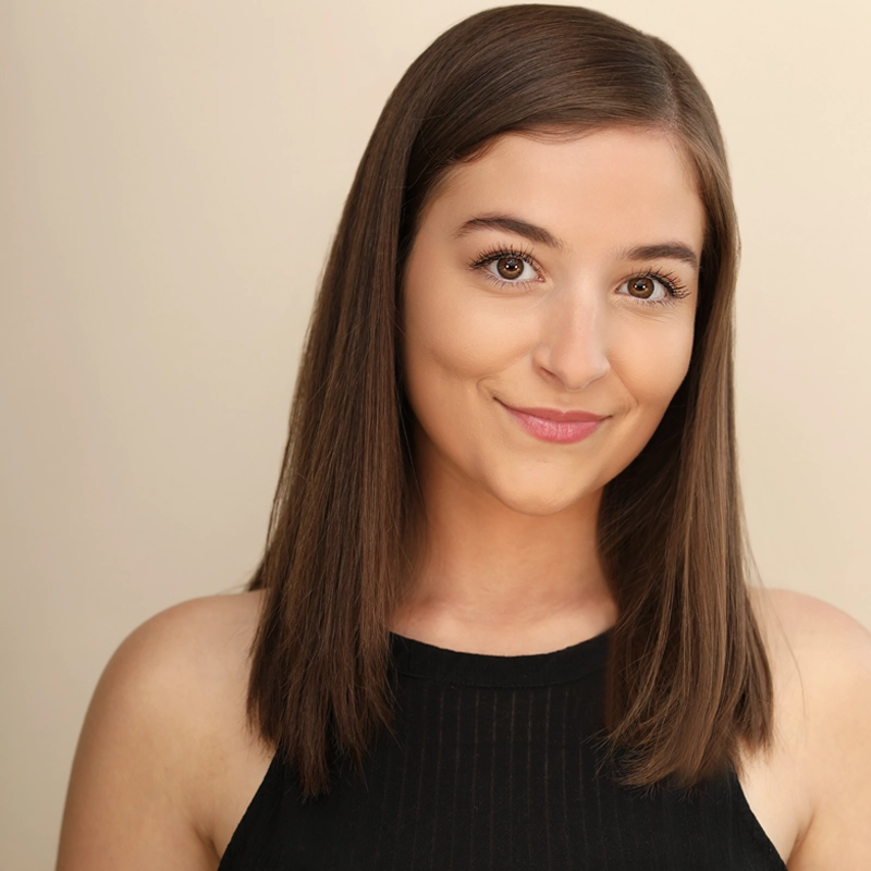 A young woman with straight, shoulder-length brown hair and light skin smiles gently. Isabel Hartzell wears a sleeveless black top and stands against a plain beige background, looking directly at the camera with a relaxed, confident expression.