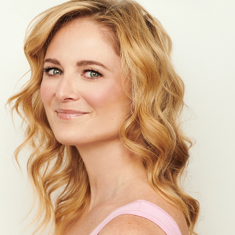 A woman with wavy, shoulder-length blonde hair smiles softly, facing left in a close-up portrait. She has fair skin, green eyes, and wears natural makeup. She is dressed in a light pink sleeveless top against a plain, light background.