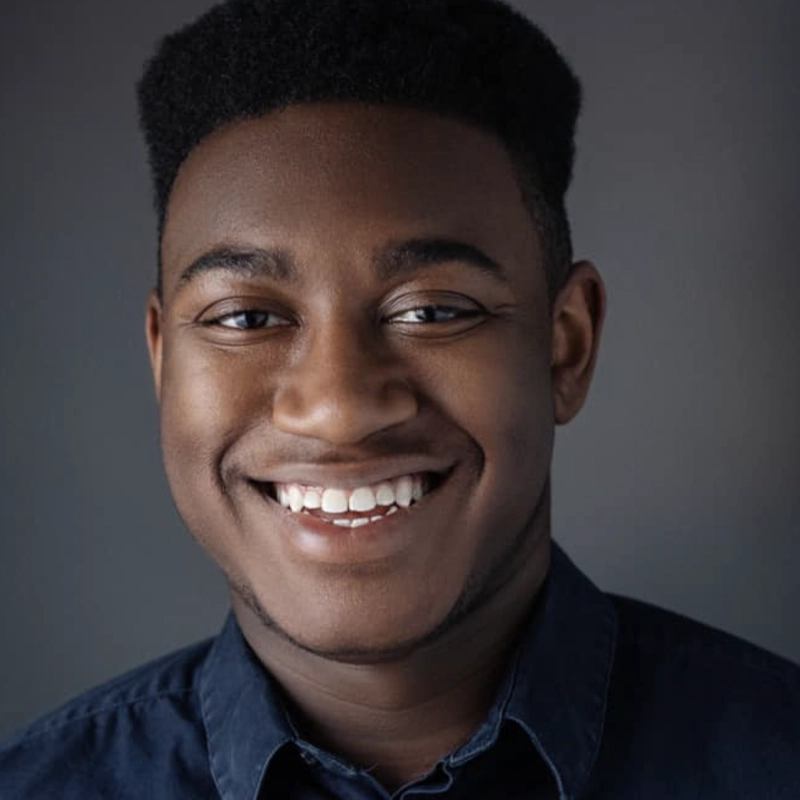 A young man with dark skin and short, styled hair smiles warmly, showing his teeth. Wesley Hodges wears a dark button-up shirt and is posed against a softly lit gray background, capturing a friendly and approachable expression.