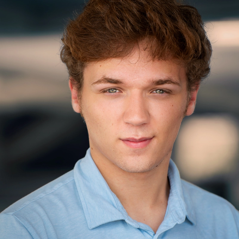 A young man with fair skin and short, wavy brown hair gazes directly at the camera, wearing a light blue collared shirt. The background is softly blurred in neutral tones, drawing focus to his face with a calm, confident expression.