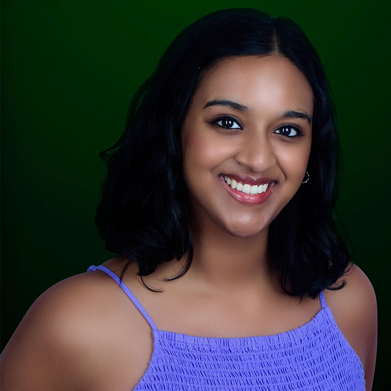 Young woman with medium skin tone and shoulder-length black hair, smiling at the camera. She wears a sleeveless lavender top with thin straps and a ruched bodice. The background is a smooth gradient of dark green. Emma Wallace style.