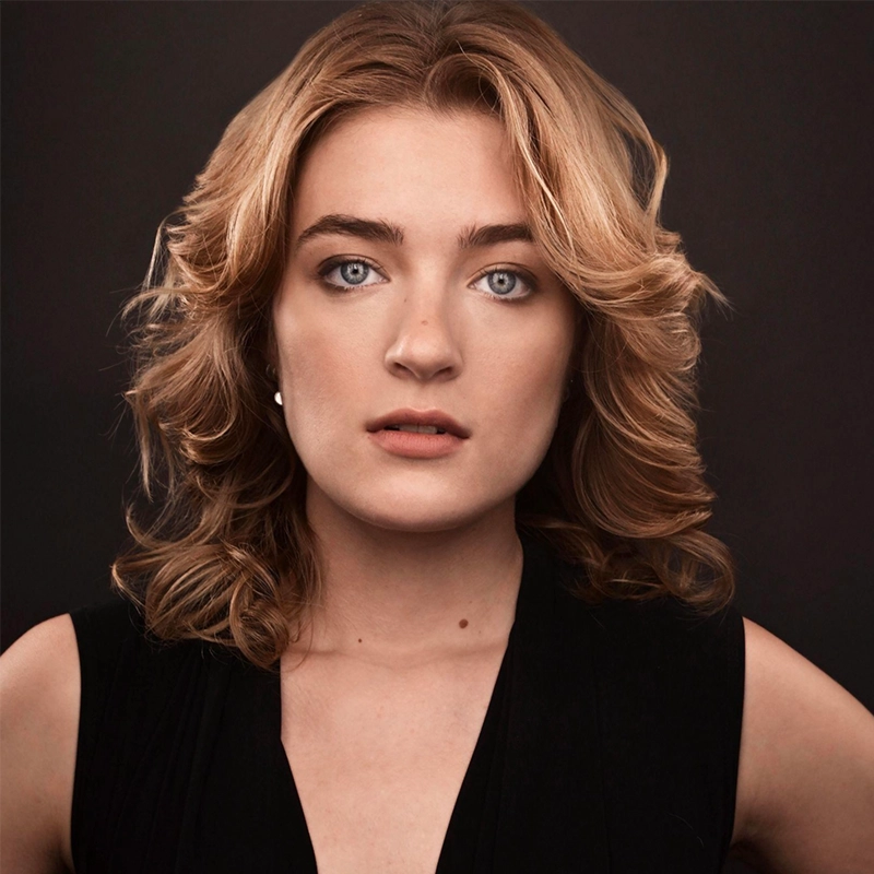 Amanda Harris, with wavy, shoulder-length blonde hair, poses against a dark background. She has blue eyes, fair skin, and wears a sleeveless black top. Her expression is neutral as she looks directly at the camera.