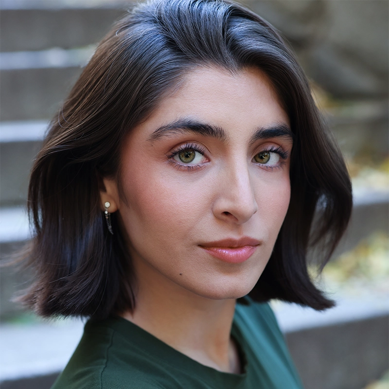Jaeden Riley Juarez, with short, dark brown hair and hazel eyes, looks directly at the camera. She wears a green top and subtle makeup. Blurred stone steps and yellow leaves in the background create a natural, outdoor setting.
