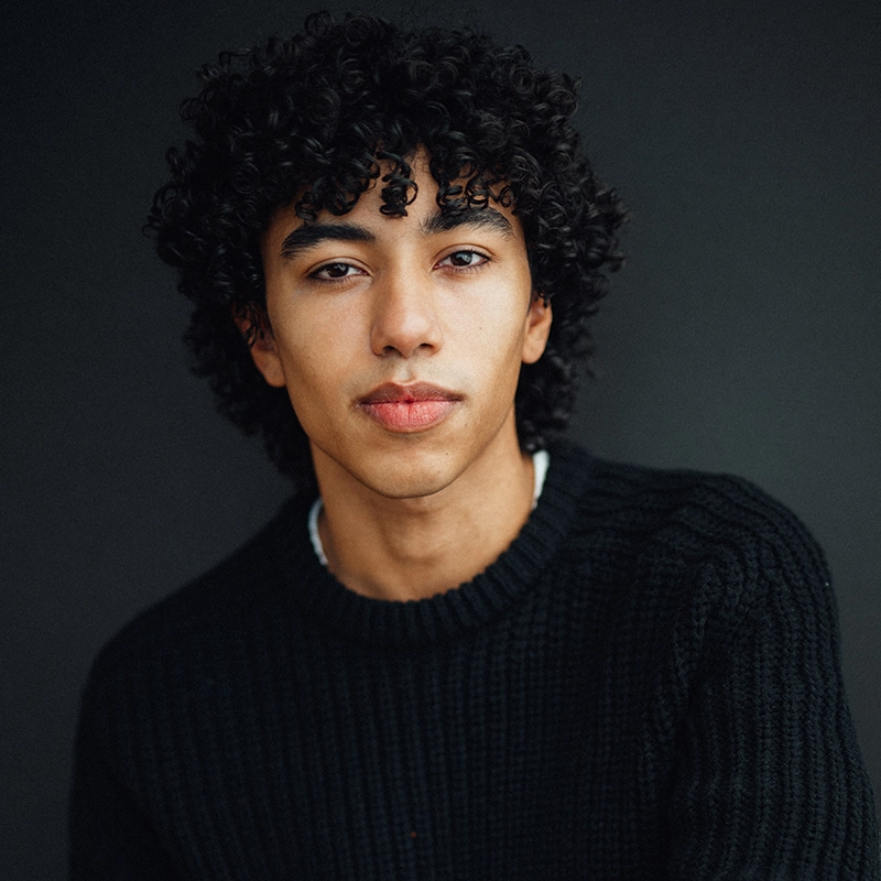 A young man with medium skin tone and short, curly black hair stares calmly at the camera. He wears a black knit sweater. The dark, neutral background highlights Sergio Guerra’s thoughtful expression and the soft lighting on his face.