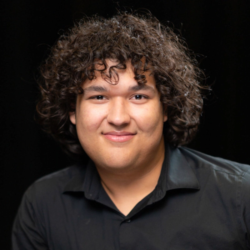 A young person with medium skin, curly brown hair, and a slight smile is wearing a black collared shirt. In this professional head-and-shoulders portrait, Sergio Guerra poses against a plain black background, looking directly at the camera.