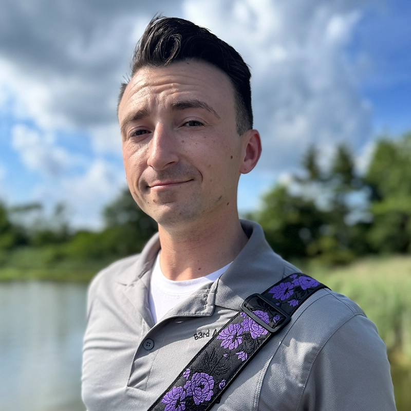 Jake McDonnell Jake McDonnell, a man with short dark hair, stands outdoors by water, wearing a light gray polo shirt and carrying a bag with a purple floral strap. Trees and a cloudy blue sky are in the background under bright, natural sunlight.