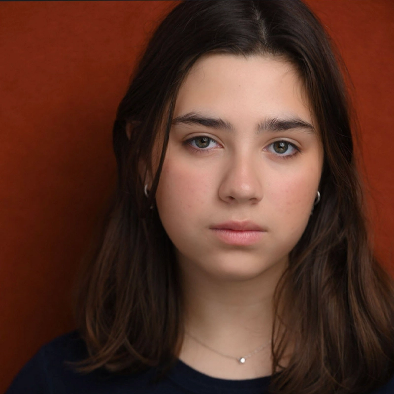Sofia Jaramillo, a young person with straight, medium-length brown hair and fair skin, looks directly at the camera. They wear a dark top, a thin necklace, and small hoop earrings against a solid reddish-brown background. Their expression is neutral.