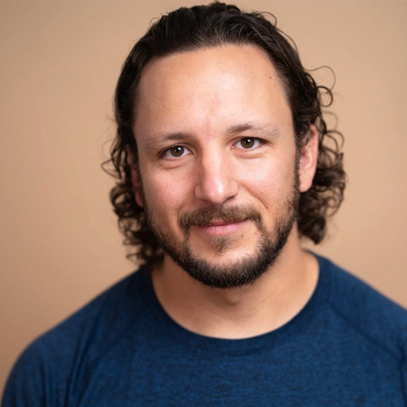 Jason Shaffer, a man with light skin, medium-length curly brown hair, and a short beard, smiles gently. He wears a dark blue crew-neck shirt and stands in front of a plain beige background with soft, even lighting.
