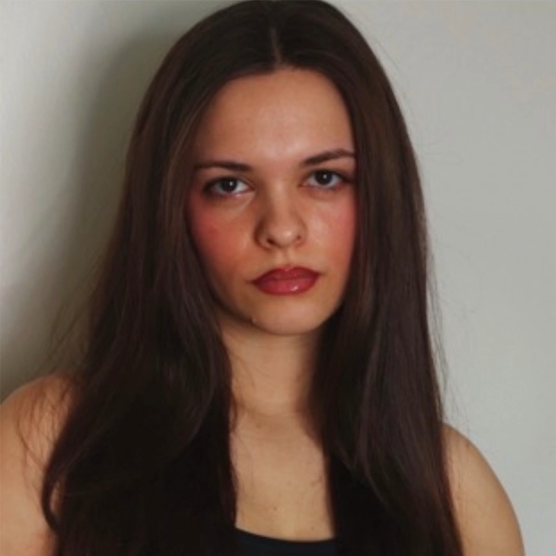 Olivia Luzquinos stands against a plain light background, her long dark brown hair straight and sleek. With medium skin, brown eyes, and natural rosy makeup, she wears a dark sleeveless top and maintains a neutral expression.