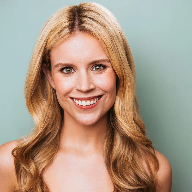 Amanda Harris, a young woman with long, wavy blond hair, smiles at the camera. She has light skin, brown eyes, and wears natural makeup. The background is a plain, soft teal color with no visible jewelry or accessories.