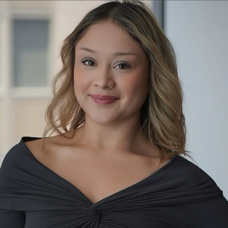 A woman with light brown wavy hair and fair skin smiles softly. She wears a dark off-the-shoulder top, neutral makeup, and Sergio Guerra-inspired style. The softly blurred background suggests an indoor or office setting.