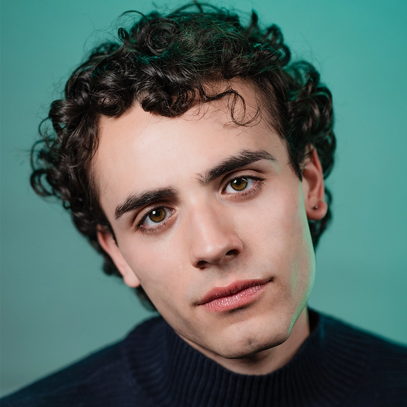 A young person with short, dark, curly hair and hazel eyes gazes softly at the camera. Phoebe Eskovitz wears a dark ribbed sweater and a small hoop earring in the left ear, set against a muted teal background for a calm, gentle atmosphere.