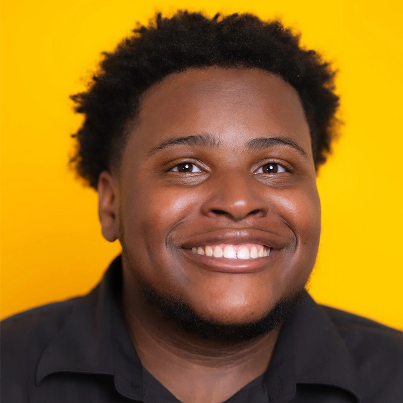 Josiah Jennings, a young man with short curly hair and a trimmed beard, smiles broadly, showing his teeth. He wears a black collared shirt and stands against a bright yellow background, the even lighting highlighting his cheerful expression.