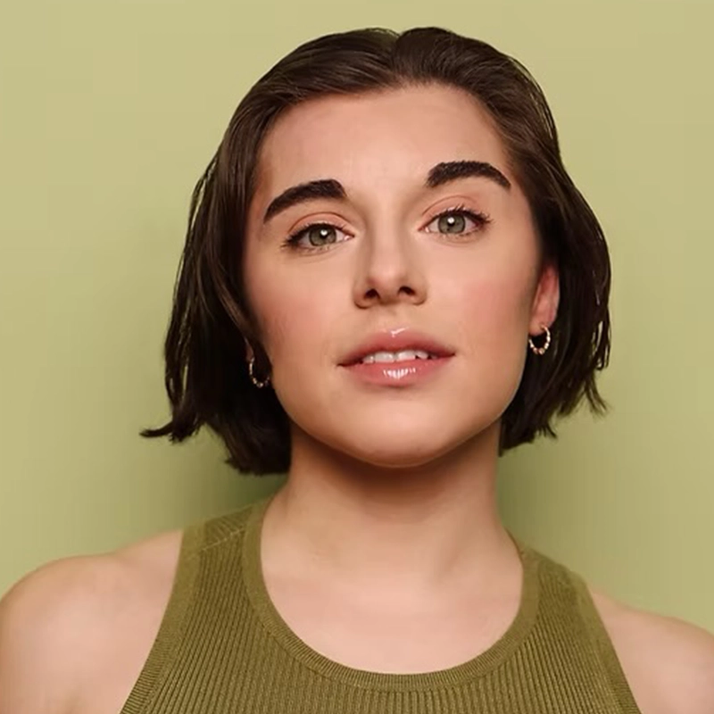 Adriana Ellis, with short, dark brown hair and light skin, stands against a plain, light olive-green background. She wears a green sleeveless top and small hoop earrings. Her expression is neutral with a slight smile and natural makeup.