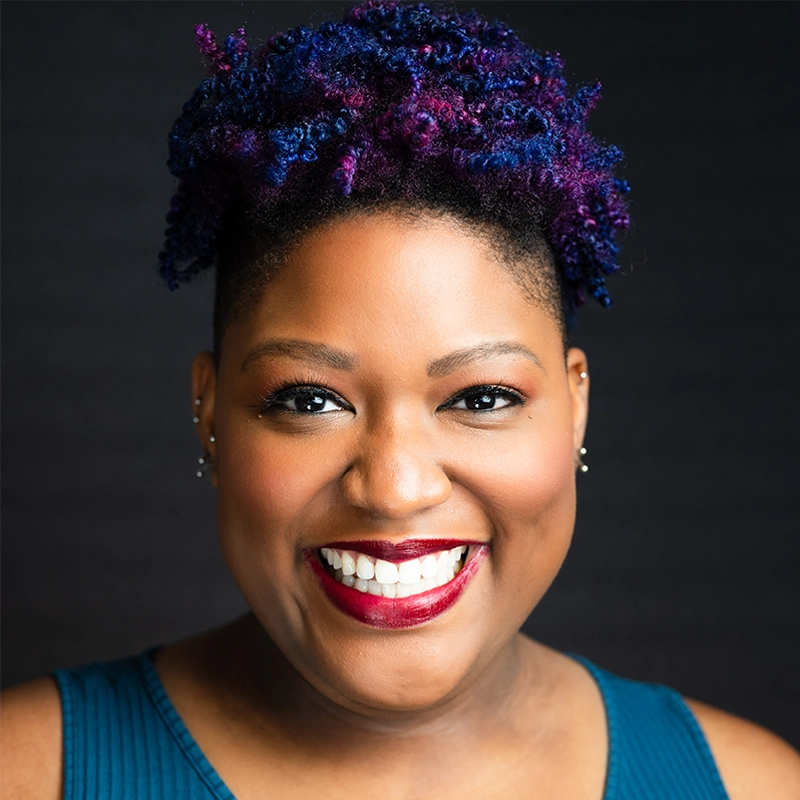 A smiling woman with deep skin tone and bright purple and blue curly hair styled up. She wears stud earrings, bold red lipstick, and a teal sleeveless top. Evan Swanson captures her cheerful expression and vibrant features against a black background.