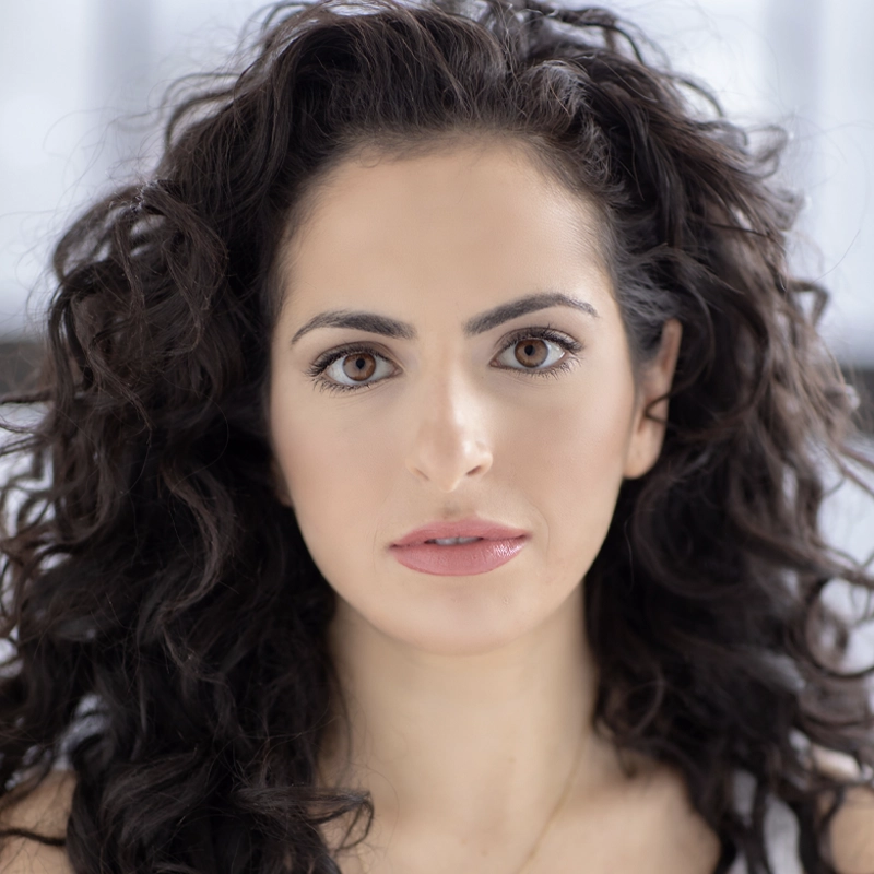 A woman with long, thick, dark curly hair and light skin looks directly at the camera. She has brown eyes, well-groomed eyebrows, subtle pink lipstick, and wears a white top. The background is softly blurred in light tones.