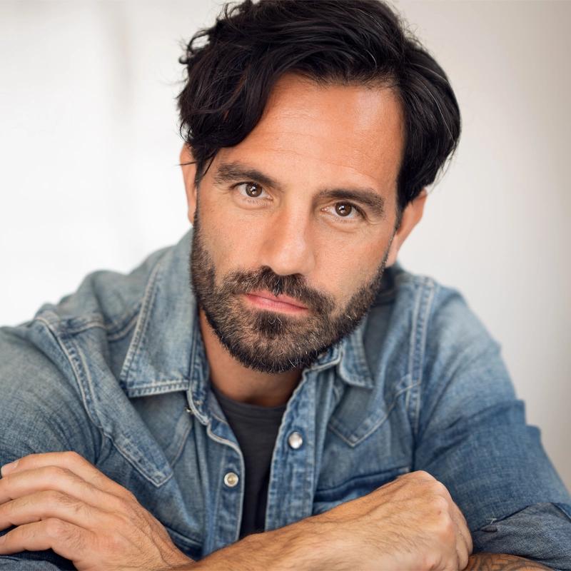 A man with wavy dark hair and a full beard, resembling Ramin Karimloo, looks directly at the camera. He wears a light denim button-up shirt over a dark t-shirt, resting his arms on a surface in front of him. The background is softly blurred and neutral.