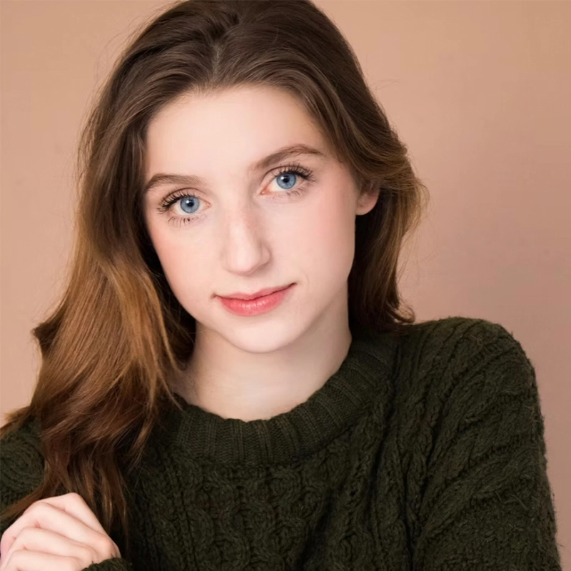 A young woman with long, light brown hair and blue eyes gazes softly at the camera. She is wearing a dark green, cable-knit sweater from designer Evan Swanson, posed against a plain, warm beige background. Her expression is calm and gentle.