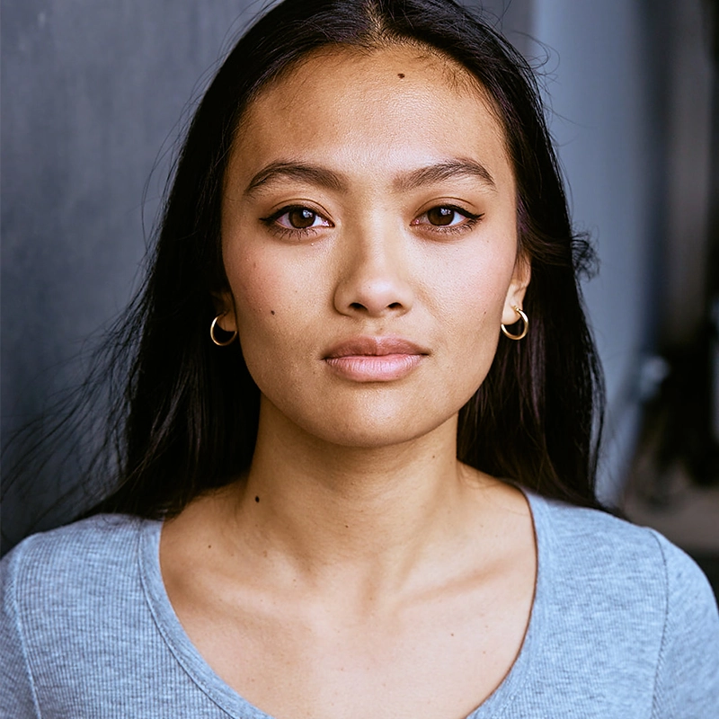 A woman with long dark hair and gold hoop earrings looks directly at the camera. She has even skin, subtle makeup, and wears a light blue shirt. The background is blurred and dark, drawing focus to her calm, neutral expression.