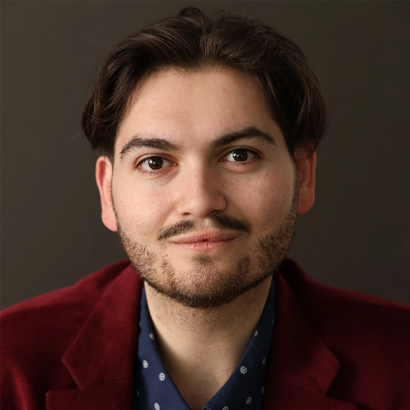 A young man, Evan Swanson, with medium-length dark hair and a trimmed beard is smiling softly. He wears a maroon blazer over a navy blue shirt with small white patterns. The plain, dark background highlights his face.