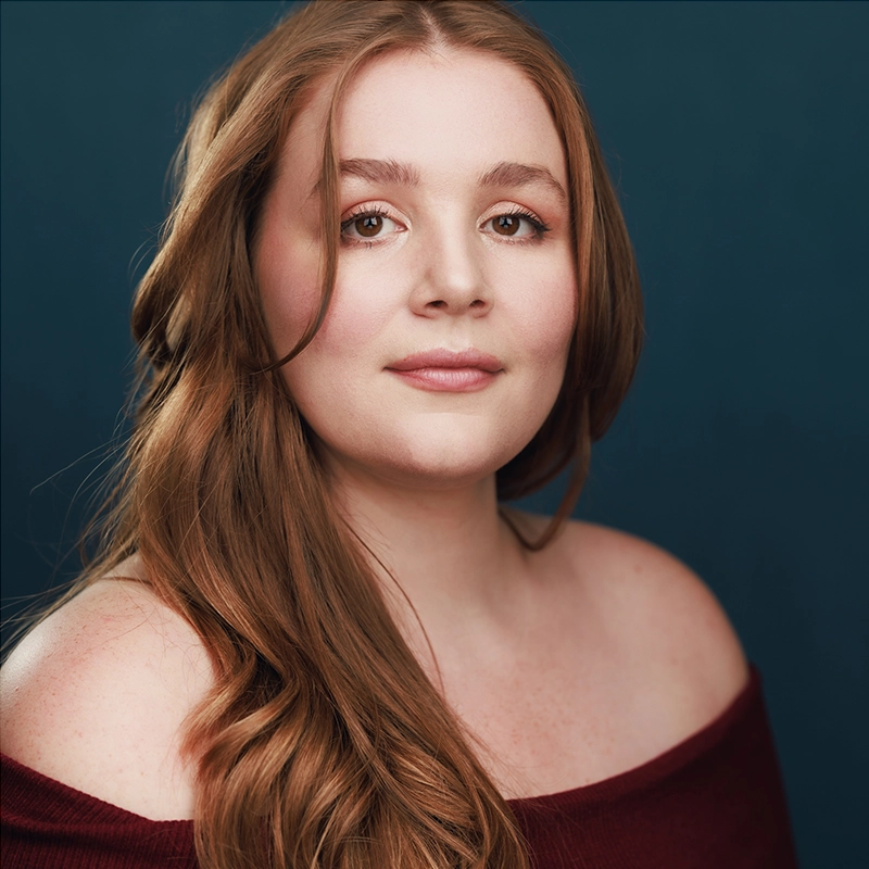 Adriana Ellis, a young woman with long, wavy auburn hair, poses in front of a dark blue background. She has fair skin, natural makeup, and wears an off-the-shoulder burgundy top, looking directly at the camera with a calm, confident expression.