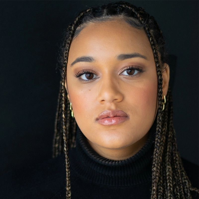 A young woman with long, braided hair looks directly at the camera against a black background. Styled by Josiah Jennings, she wears a black turtleneck, subtle makeup, and gold hoop earrings. Her expression is calm and composed.