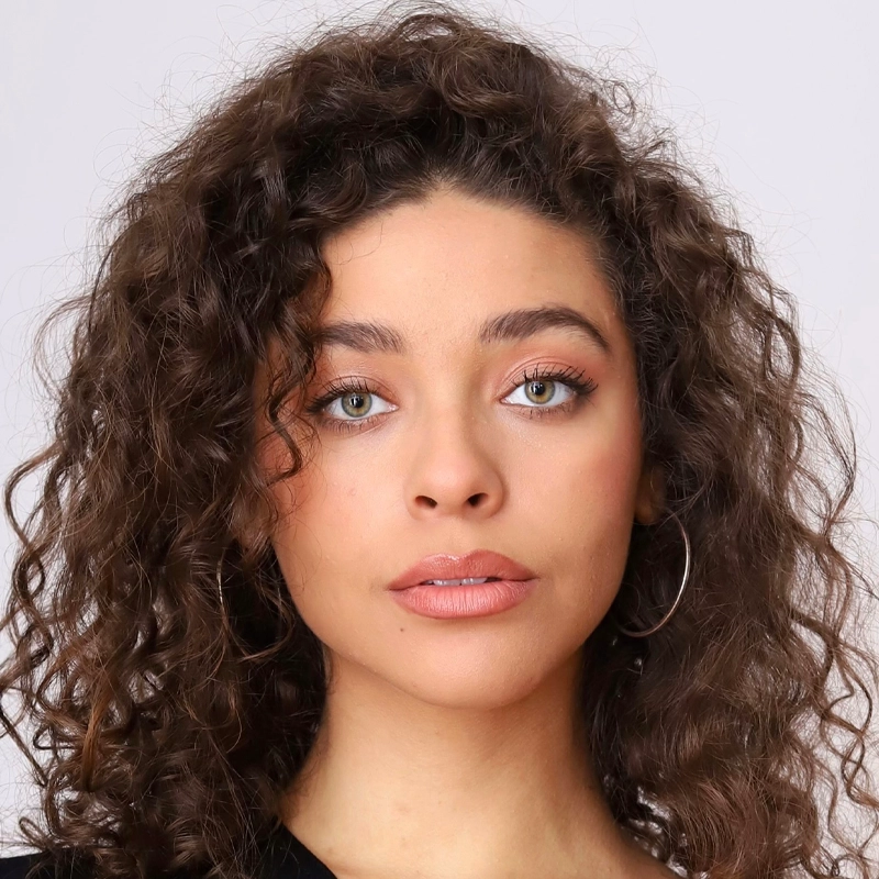 A young woman with light skin and curly brown hair looks directly at the camera. She has light-colored eyes, natural makeup, and wears hoop earrings. Her expression is neutral, and she is dressed in a black top against a plain light background.