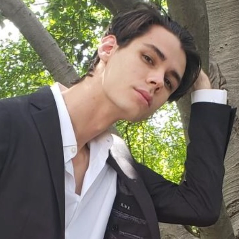 A young man with short dark hair, wearing a black suit jacket over a white shirt, leans against a tree outdoors. Kris Kusnierz gazes at the camera with a neutral expression as sunlight filters through green leaves in the serene scene.