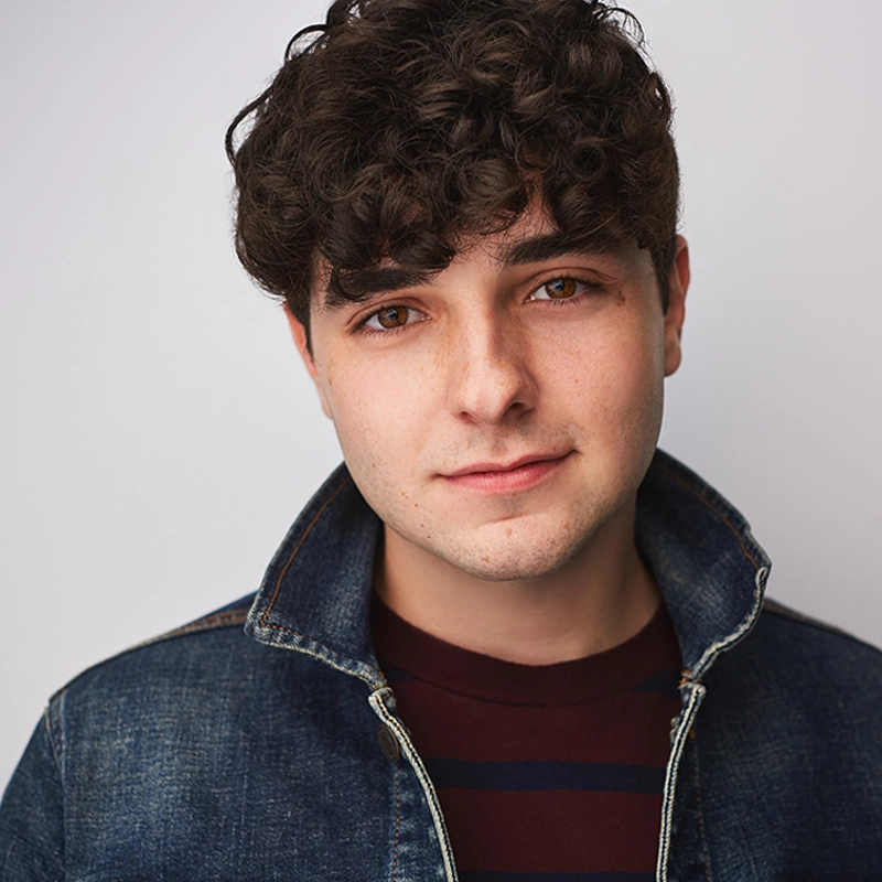 A young man with curly brown hair and fair skin looks at the camera. Kyle McArthur wears a dark denim jacket with the collar flipped up over a maroon and black striped shirt. The background is plain and light-colored. He has a slight, friendly smile.