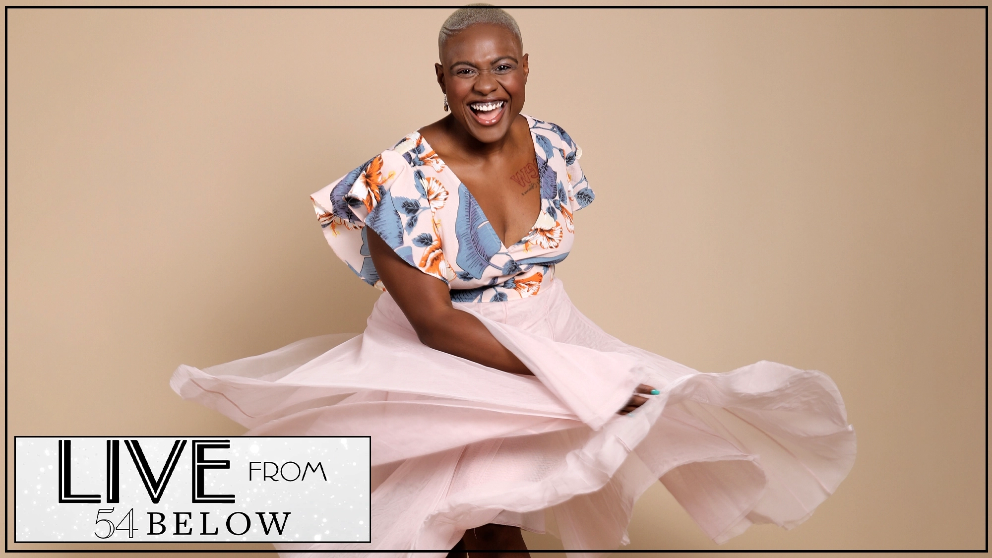 A person with short hair smiles joyfully while twirling in a soft pink skirt and floral ruffled blouse. The beige background sets the scene for Nyla Watson's 