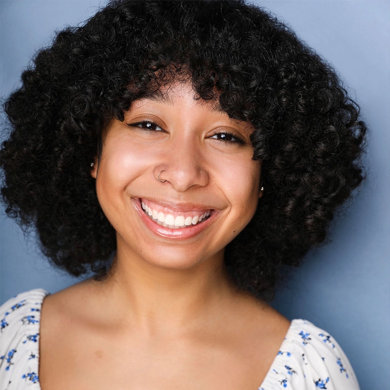 Emma Wallace, a young woman with curly dark hair, smiles warmly at the camera. She has a nose ring and wears a white blouse with blue floral patterns. The soft blue background and bright lighting highlight her friendly expression.