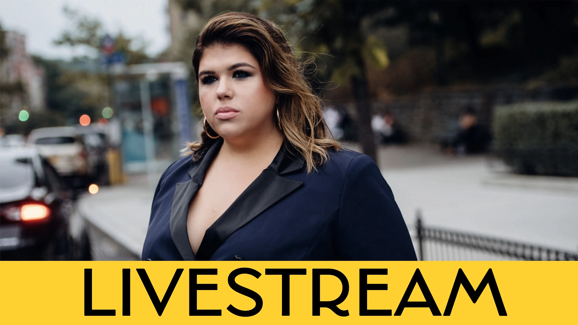 A serious woman with wavy brown hair, wearing a navy blazer, stands outdoors on a city street. Blurred cars and people are in the background. Bold black text on a yellow band reads, “LIVESTREAM: Leah Crocetto—A Quiet Thing.”.