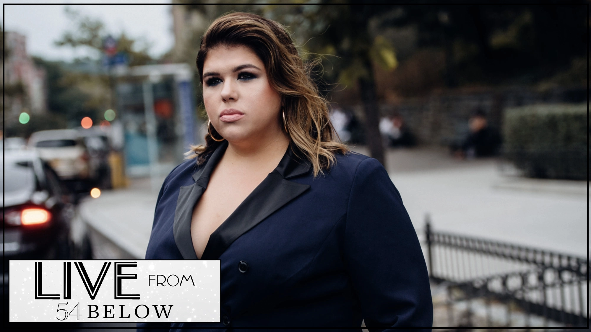 A woman with long brown hair, wearing a navy blazer with black lapels, stands outdoors on a city sidewalk. She looks ahead with a serious expression. 