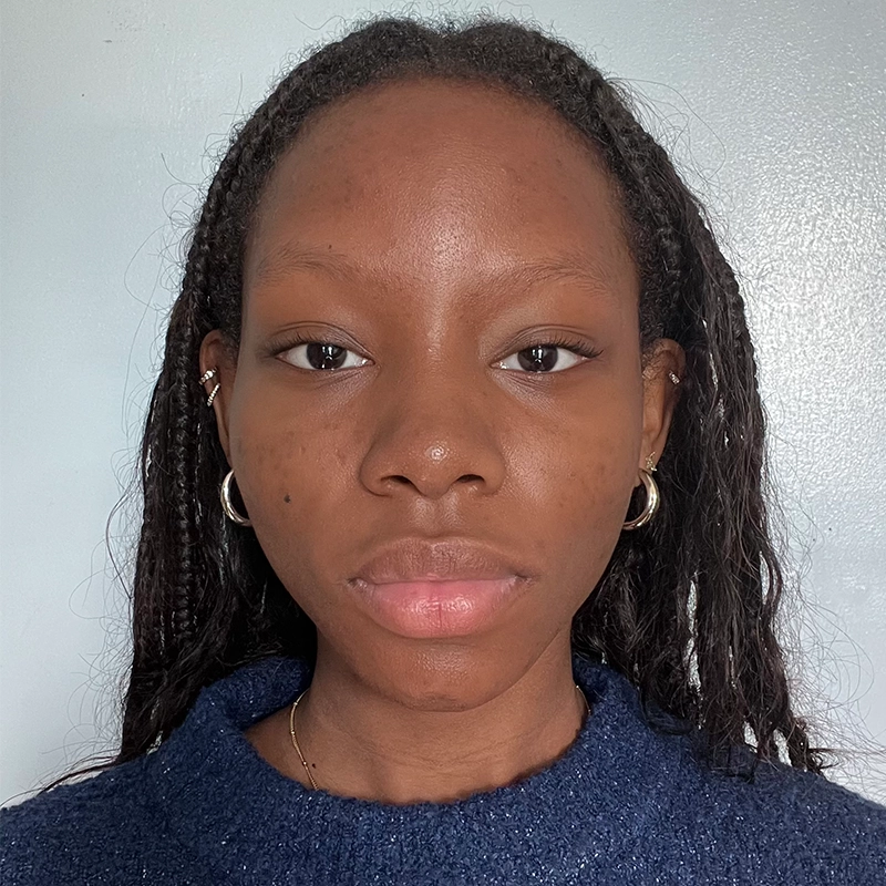 Emma Wallace, a young woman with dark skin and braided hair, stares directly at the camera against a plain light gray background. She wears a navy blue sweater, small hoop earrings, and a subtle gold necklace. Her expression is neutral.