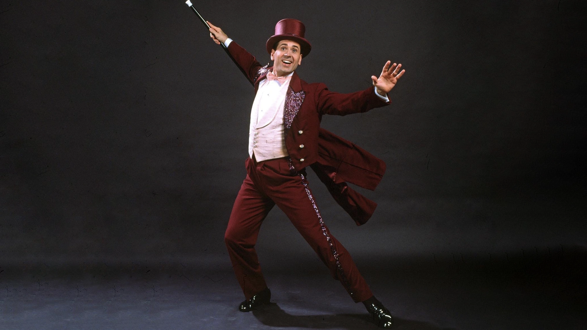 A cheerful Lee Roy Reams in a maroon tailcoat, matching top hat, white shirt, and pink vest poses energetically against a dark background—holding a black cane aloft and spreading his arms wide with a big smile, channeling the spirit of 42nd Street.